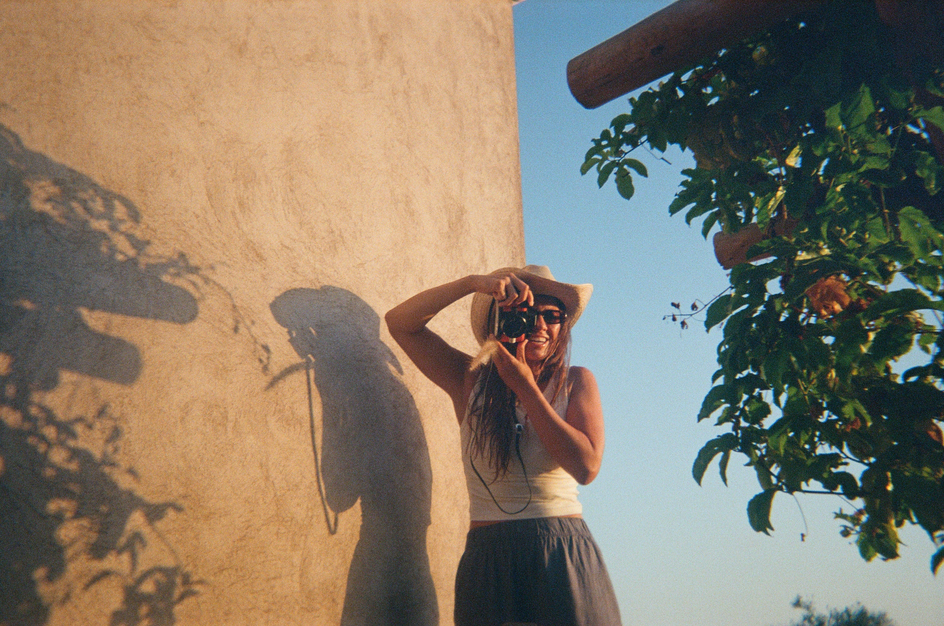 A woman wearing a wide-brimmed straw hat, sunglasses, a white tank top, and a dark skirt takes a photo with a camera against a warm beige stucco wall under a blue sky, with leafy green vines on the right.