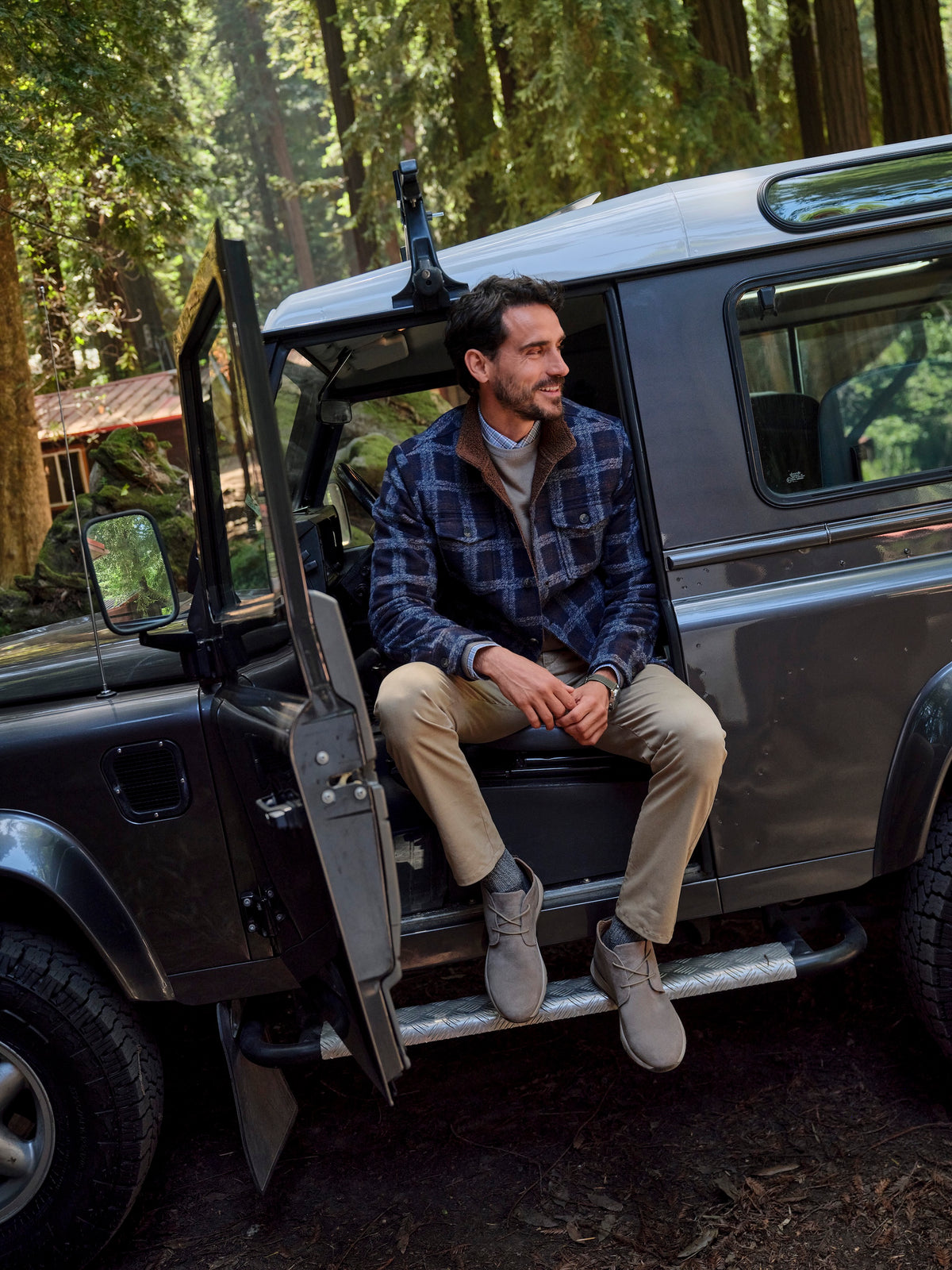 A man sits on the open step of a dark gray off-road vehicle in a forest setting, wearing a blue and black plaid flannel shirt, tan trousers, and gray boots.