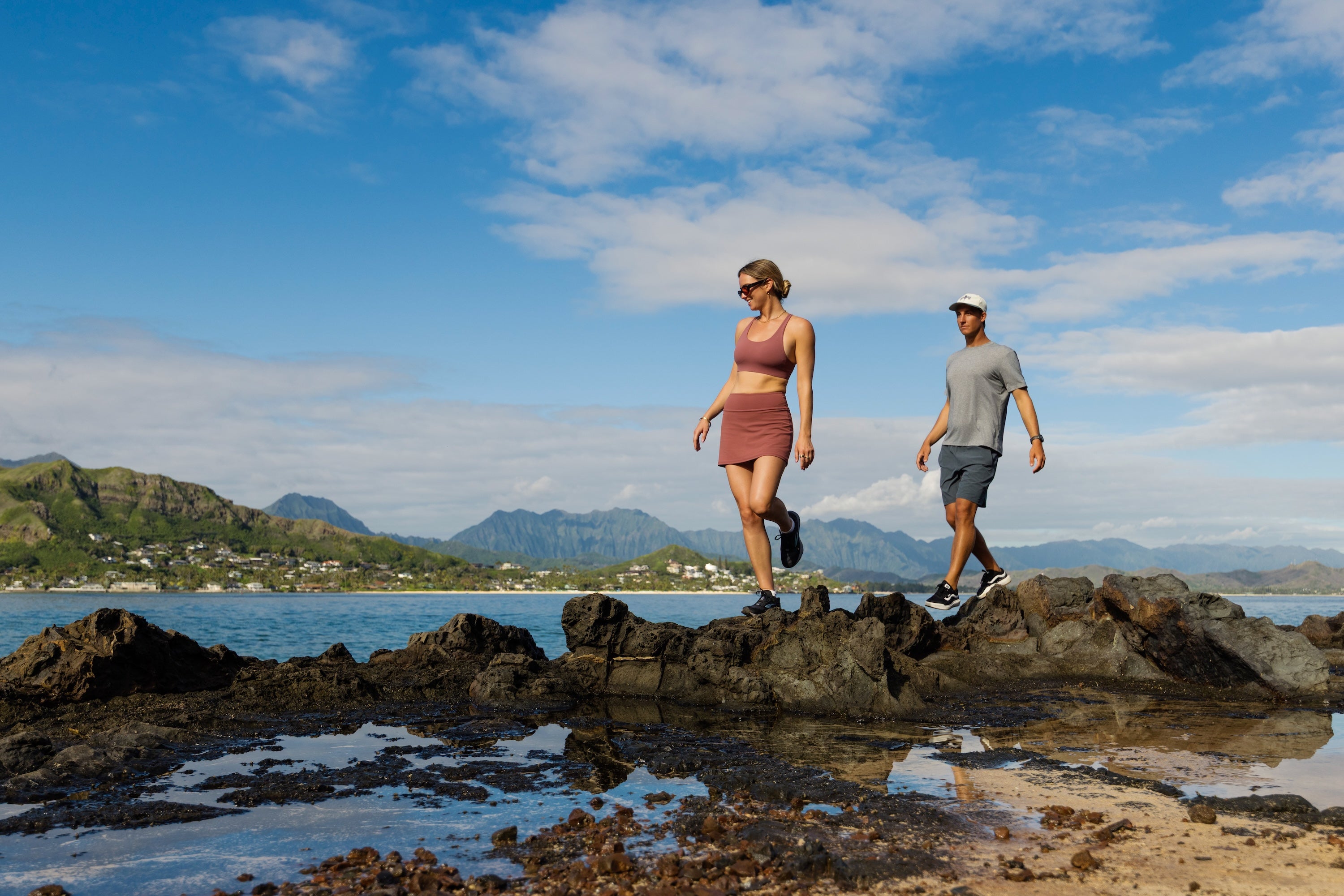 A man and a woman walking along dark rocks by the ocean under a blue sky with clouds, with mountains and water in the background.