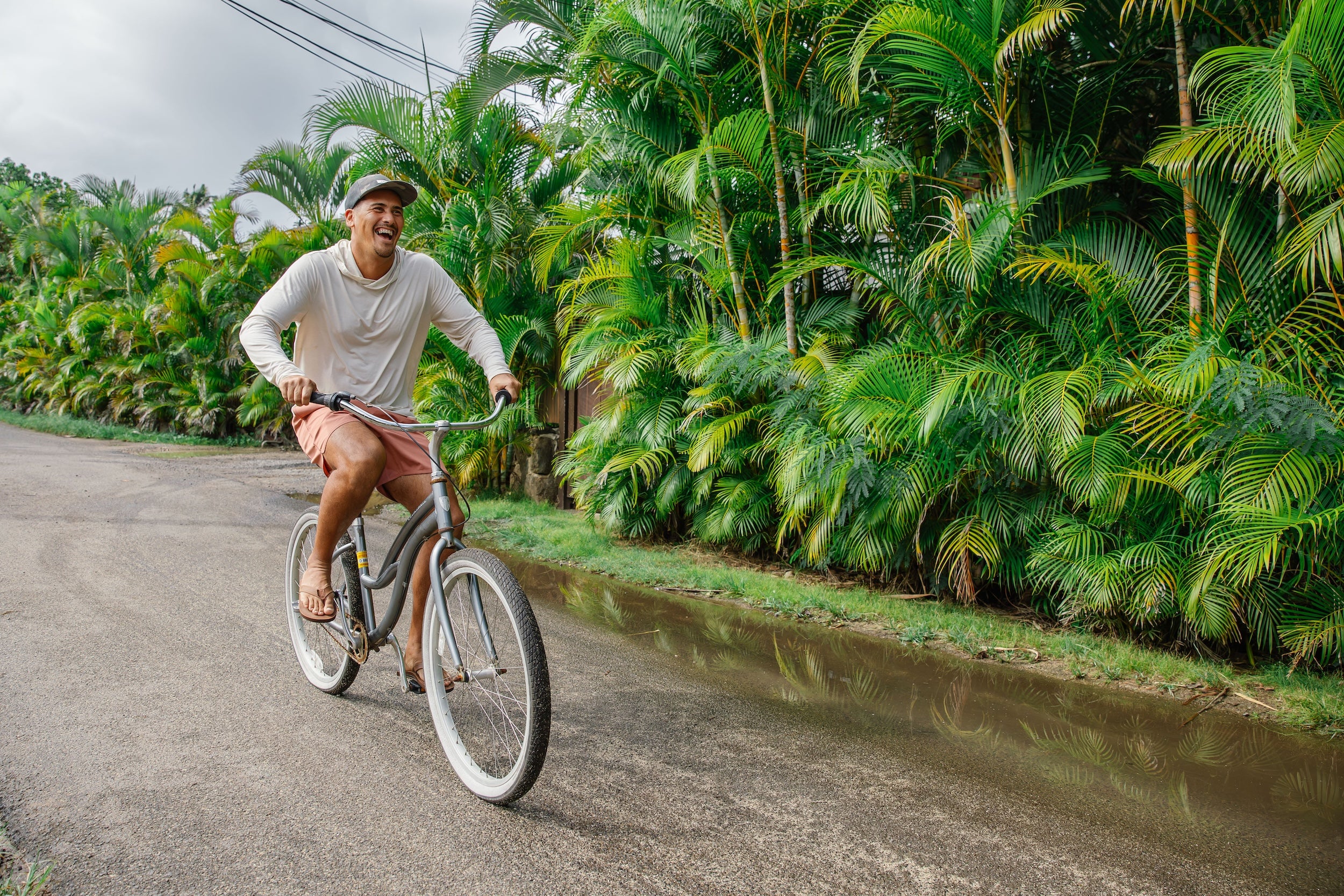 Man on bike smiling with a long sleeve hoodie on and flip flops in a tropical country