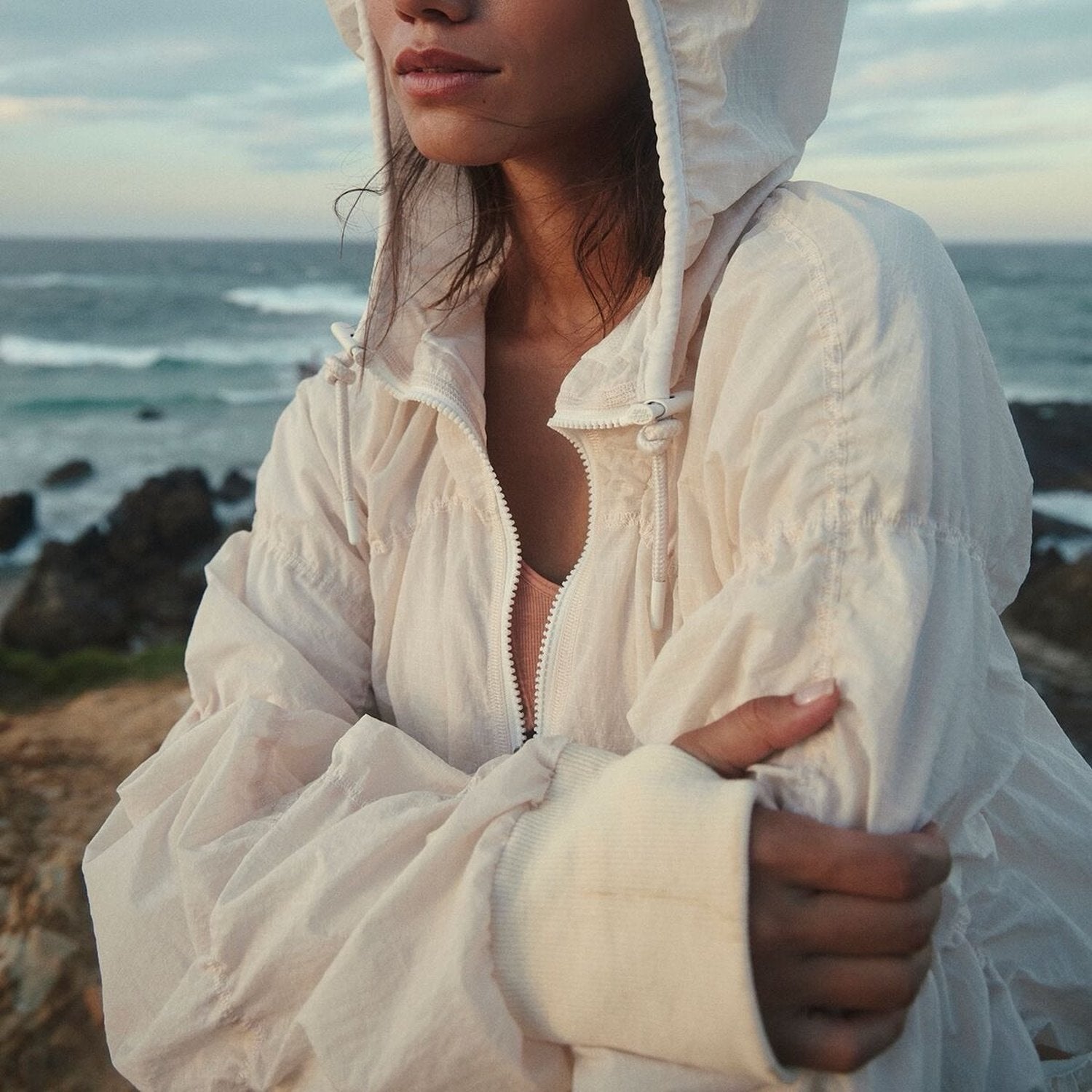 Person wearing a white hooded jacket standing on a rocky coastline with ocean waves in the background.