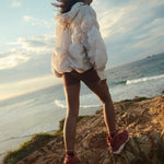 Person standing on a cliff overlooking the ocean with a white jacket and red shoes.