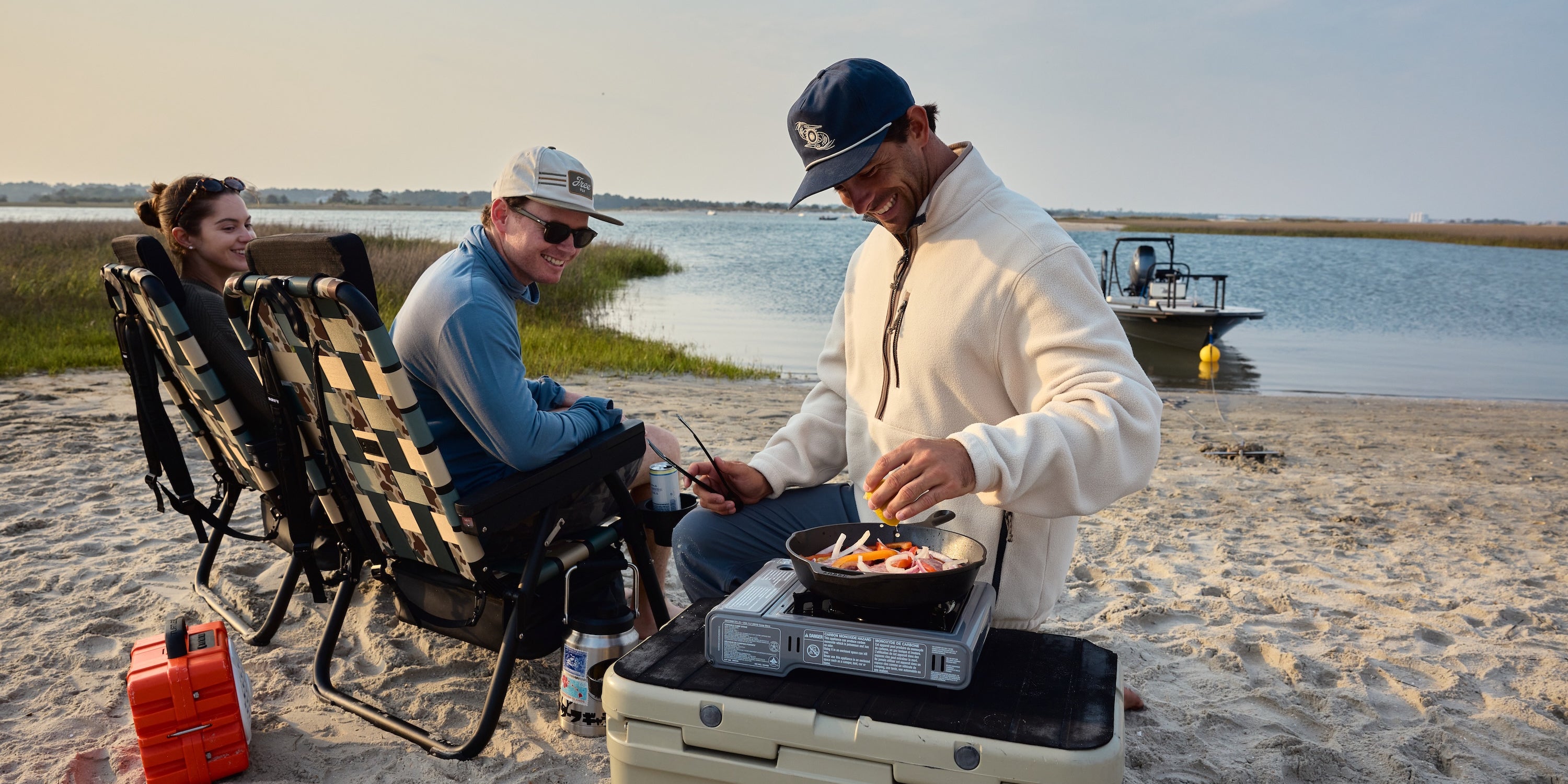 Three people camping on a sandy beach by the water, with one person cooking food on a portable stove atop a white cooler, while two others relax in black-and-white checkered lounge chairs. A red fuel canister sits nearby, and a boat is visible on the water in the background.