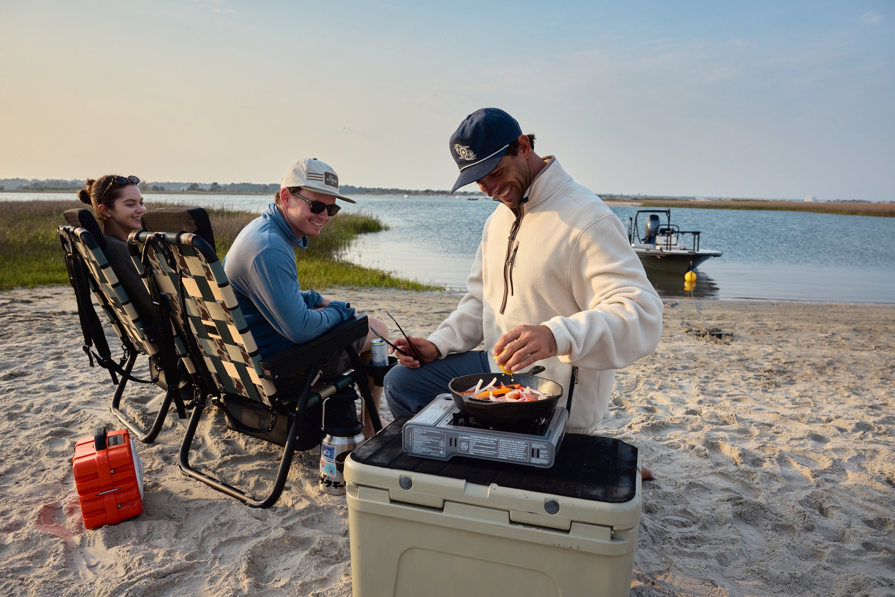 Three people camping on a sandy beach by the water, with one person cooking food on a portable stove atop a white cooler, while two others relax in black-and-white checkered lounge chairs. A red fuel canister sits nearby, and a boat is visible on the water in the background.