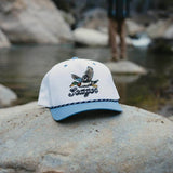 A white and light blue baseball cap with an embroidered duck and the word 'Sooner' resting on a large rock outdoors, with a blurred river and person in the background.