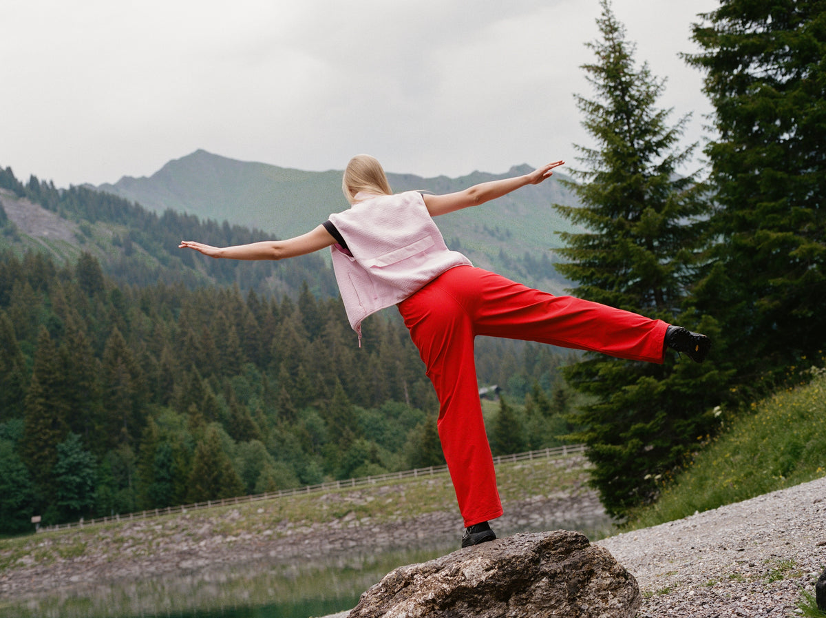 A woman posing outdoors in a mountain lake setting, wearing bright red pants, a light pink sleeveless top, and black boots while balancing on a rock.