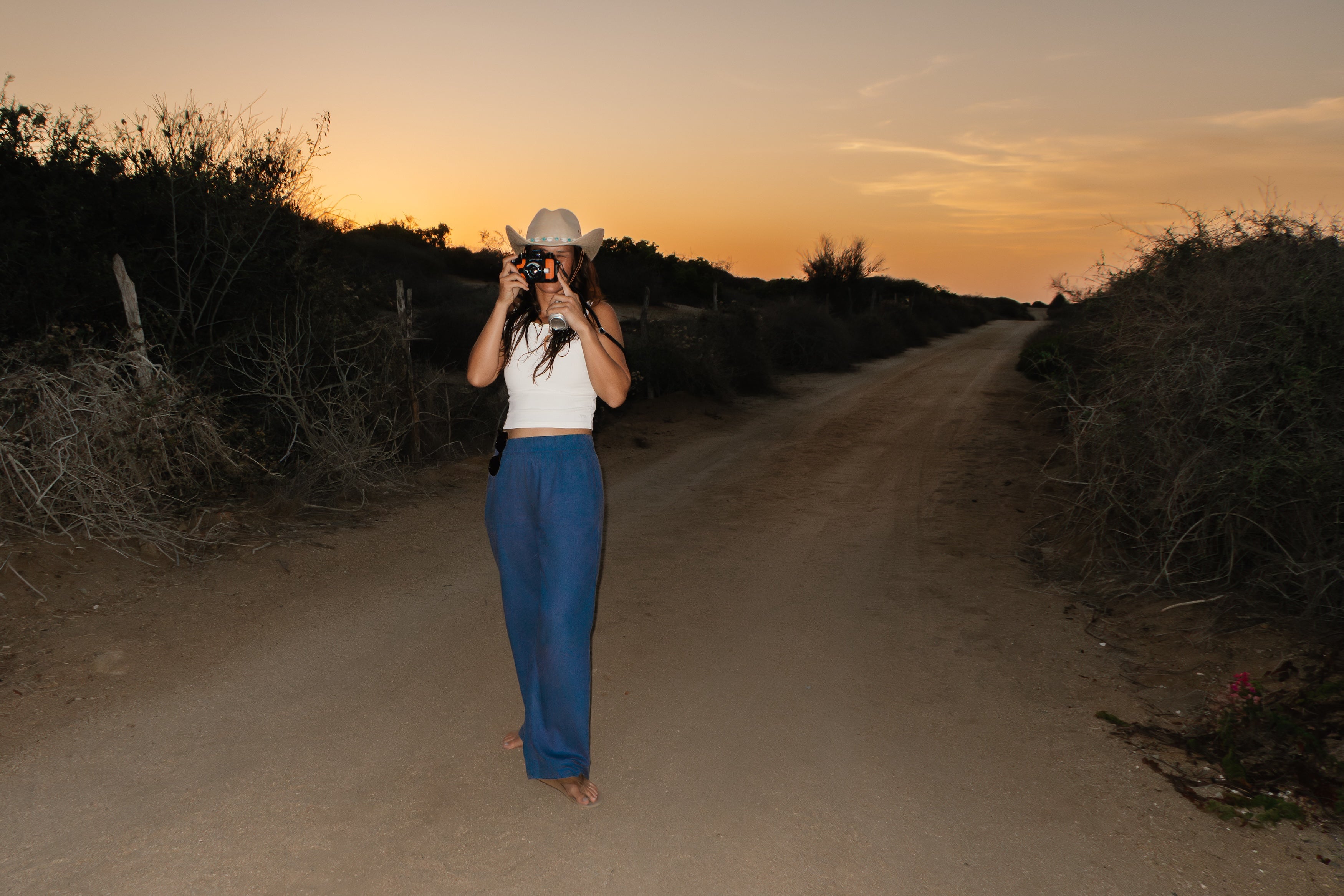 A woman wearing a white cowboy hat, white sleeveless top, and blue wide-leg pants stands on a dirt road at sunset, holding a camera up to her face. Dry brush and shrubs line both sides of the road under a warm orange sky.