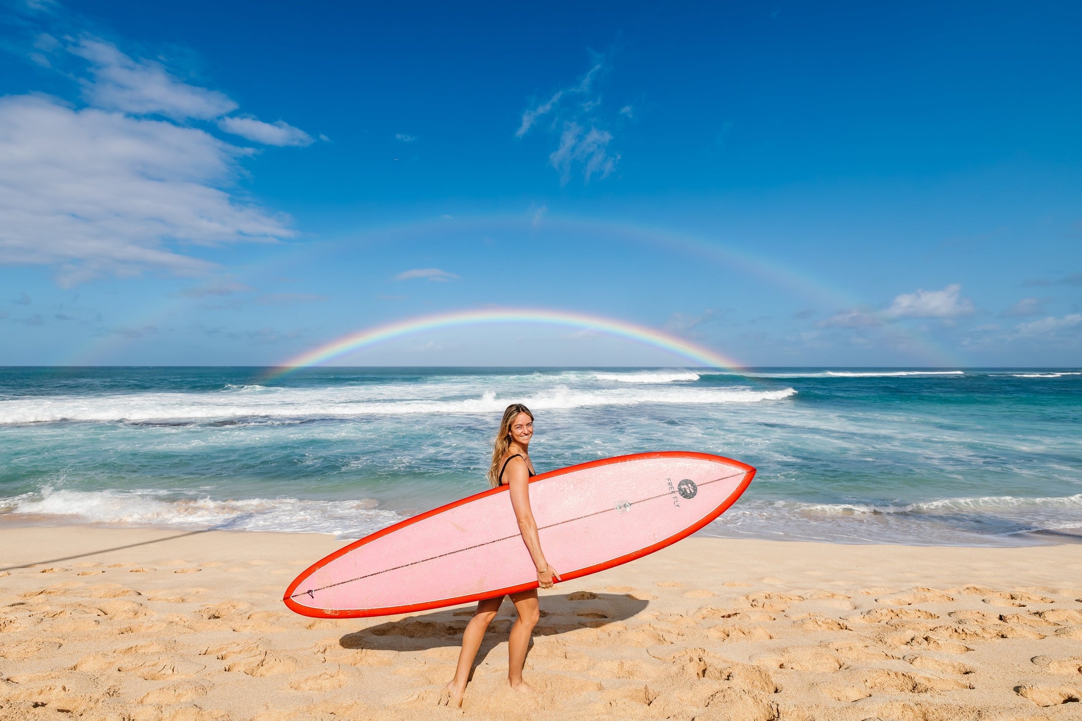 women with surfboard on beach with a rainbow over her head