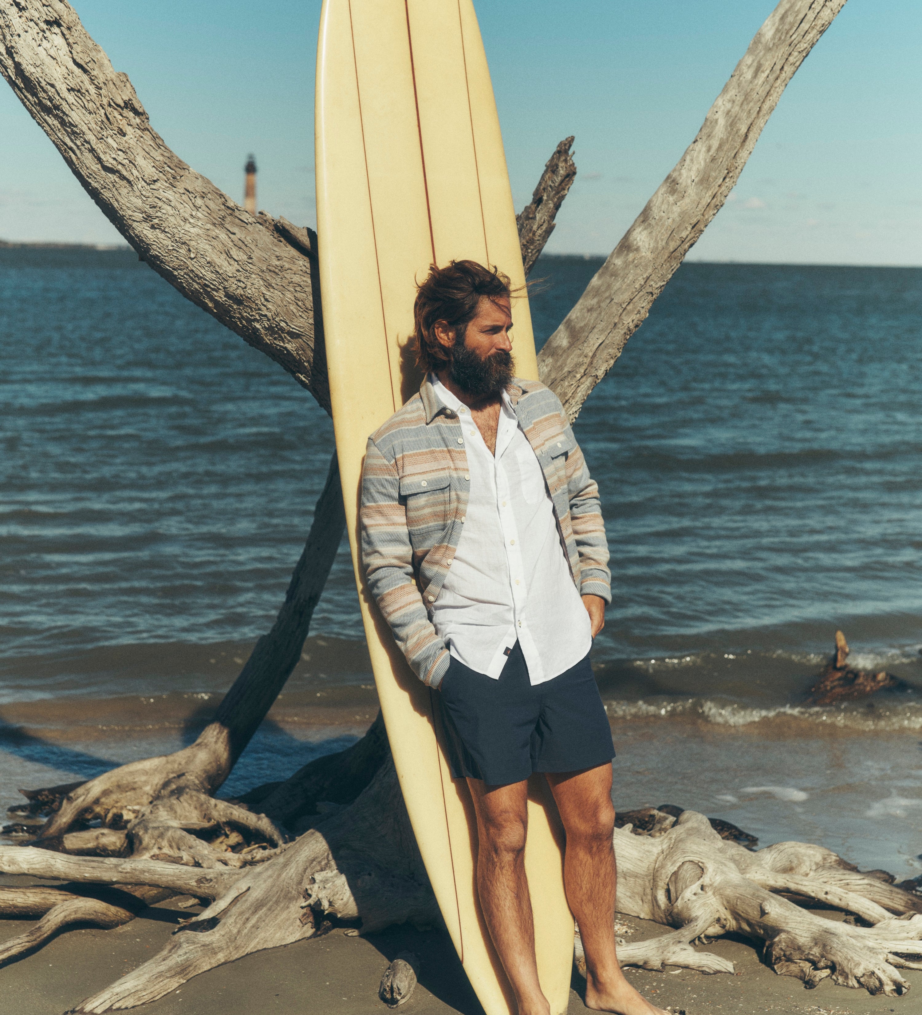 Man holding a surfboard on a beach with driftwood and ocean in the background