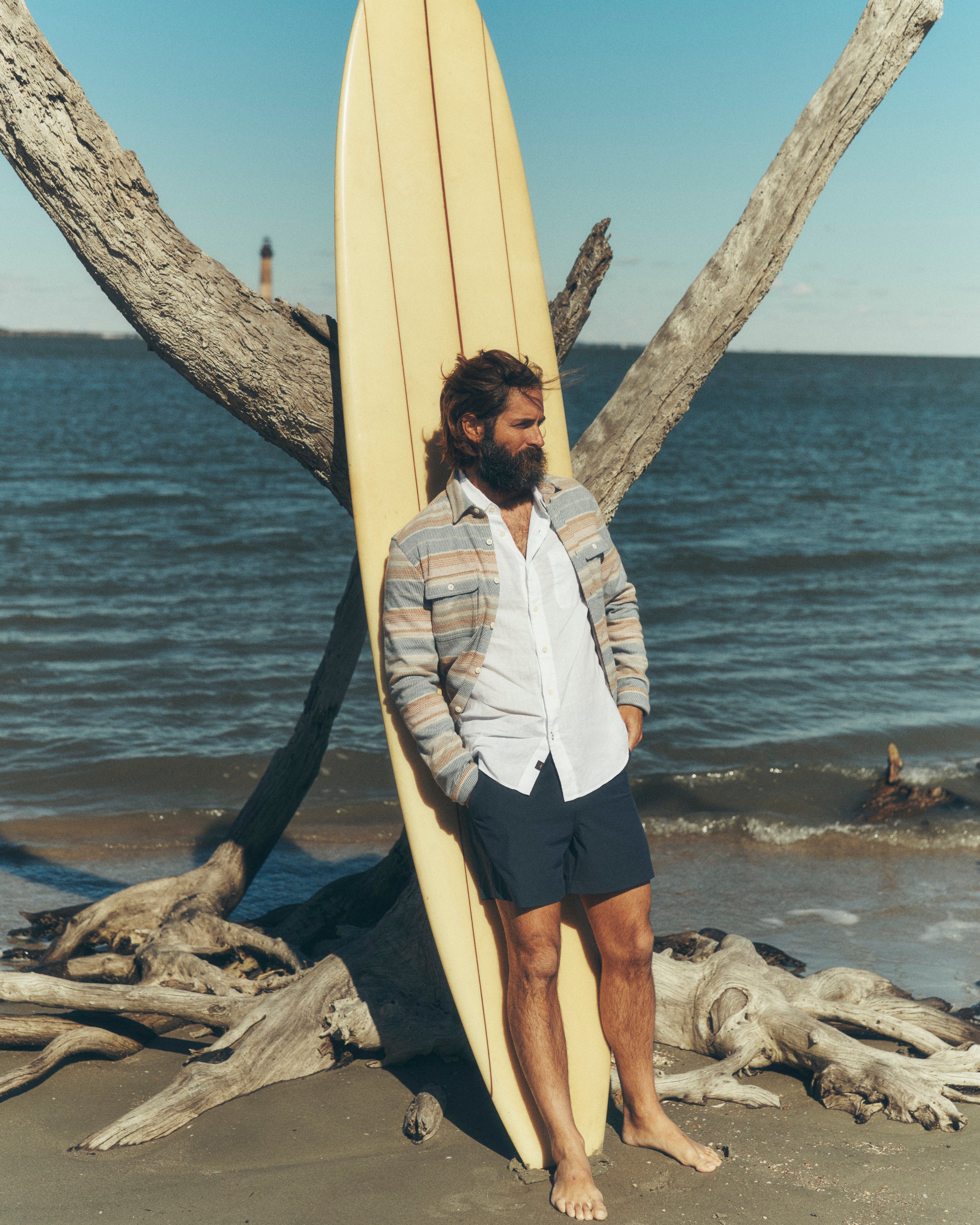 Man holding a surfboard on a beach with driftwood and ocean in the background