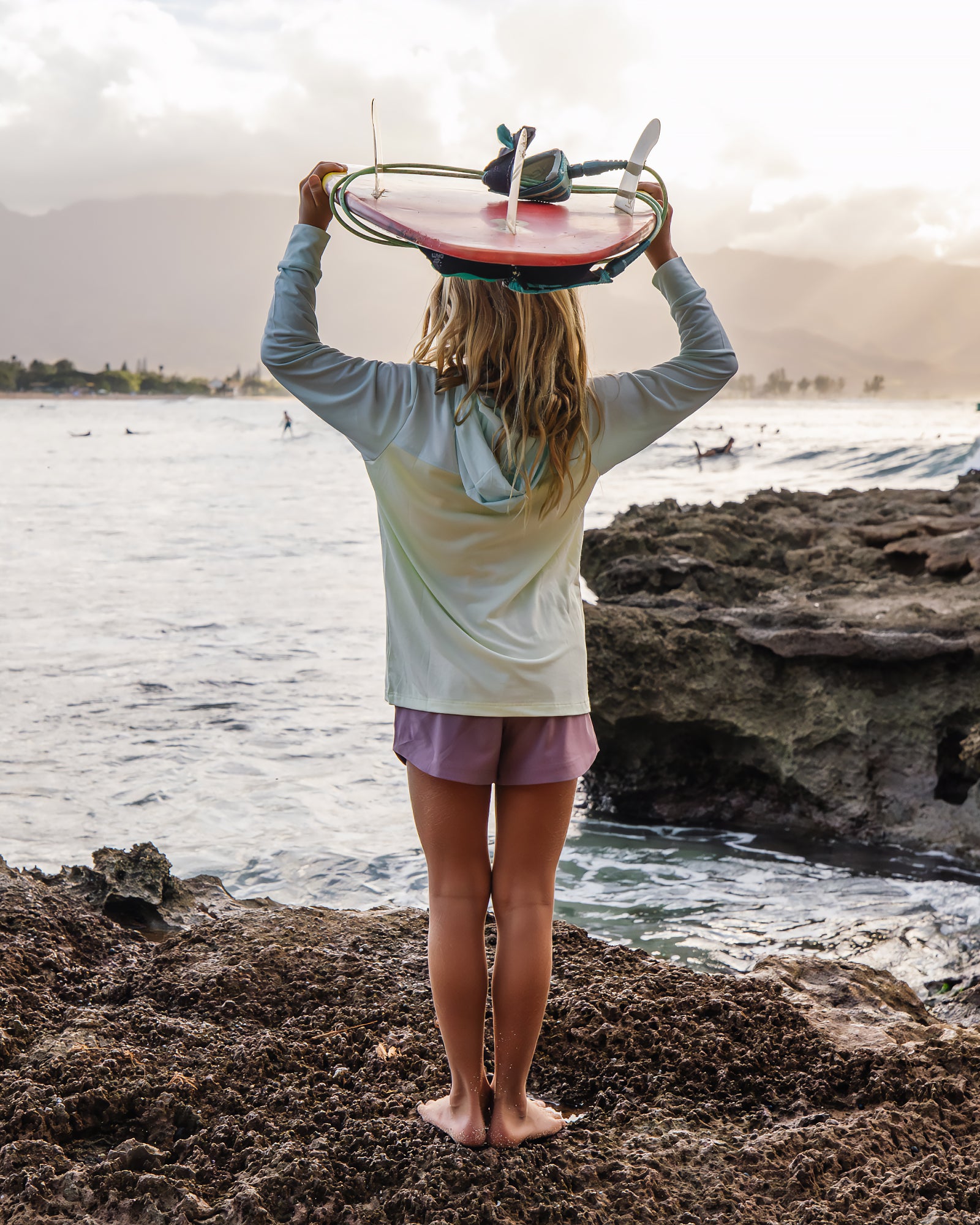 Person holding a surfboard on a rocky beach with ocean and sky in the background
