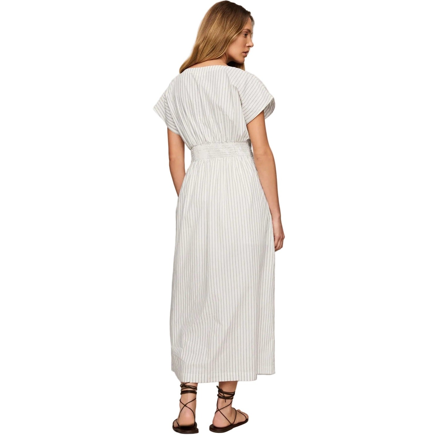 A woman shown from the back wearing a white midi dress with thin vertical gray stripes, short cap sleeves, and a matching waist belt. She is wearing dark brown strappy flat sandals against a plain light gray studio background.