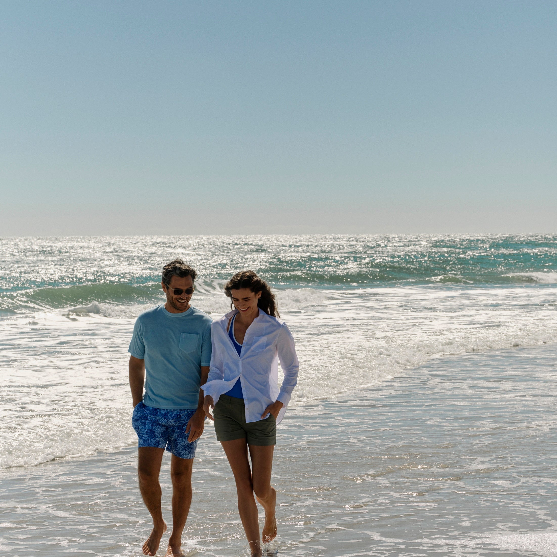A man and a woman walking hand in hand along a sandy beach with gentle waves in the background under a clear blue sky.